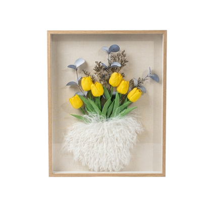 A framed floral wall art piece featuring yellow tulips with green leaves, blue-gray foliage, and a fluffy white textured base inside a light wood shadow box on a white background.