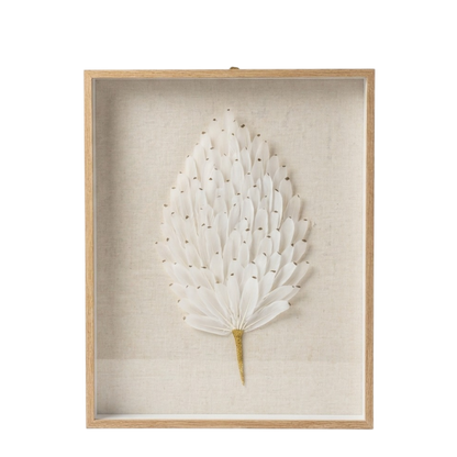 A framed botanical wall art piece featuring a white dried palm leaf with small gold accents, mounted on a textured beige linen background inside a light natural wood frame.