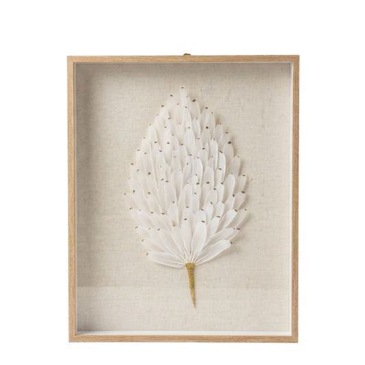 A framed botanical wall art piece featuring a white dried palm leaf with small gold accents, mounted on a textured beige linen background inside a light natural wood frame.