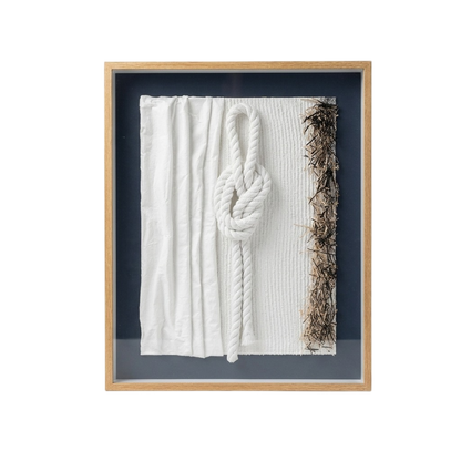 A framed wall art piece featuring a white textured fabric panel with a large white rope knot in the center and a vertical strip of brown dried seaweed or plant material along the right edge, set in a light wood frame against a dark navy background.