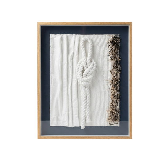 A framed wall art piece featuring a white textured fabric panel with a large white rope knot in the center and a vertical strip of brown dried seaweed or plant material along the right edge, set in a light wood frame against a dark navy background.