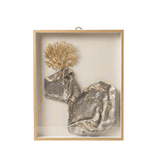 A framed abstract wall art piece with a light wood frame and clear glass cover, featuring crumpled metallic silver forms and a dried gold botanical element on a neutral off-white background.