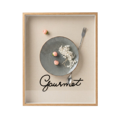 A framed wall art print featuring a minimalist food still life with a gray plate, dried white flowers, pink candy pieces, a fork, and the word 'Gourmet' in black script on a beige background, enclosed in a light wood frame.