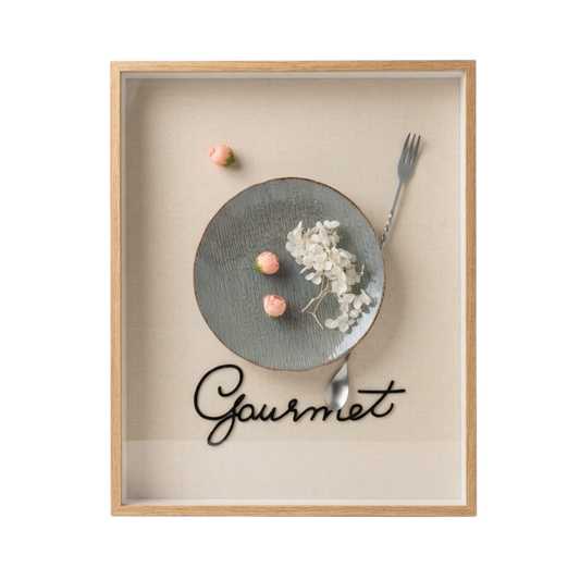 A framed wall art print featuring a minimalist food still life with a gray plate, dried white flowers, pink candy pieces, a fork, and the word 'Gourmet' in black script on a beige background, enclosed in a light wood frame.